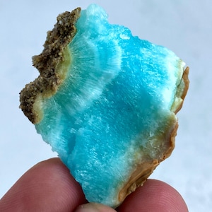 May include: A close-up of a vibrant blue mineral specimen with a layered, textured appearance. The mineral has a striking turquoise hue with white striations, and a rough, brown edge. The specimen is held in a person's hand.