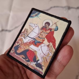 May include: Rectangular patch depicting a religious scene of a figure on horseback, spearing a dragon. The image features a haloed figure in a red cloak. The patch has a black border and is held in a hand.