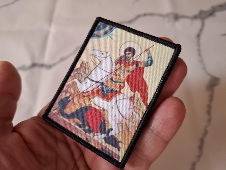 May include: Rectangular patch depicting a religious scene of a figure on horseback, spearing a dragon. The image features a haloed figure in a red cloak. The patch has a black border and is held in a hand.
