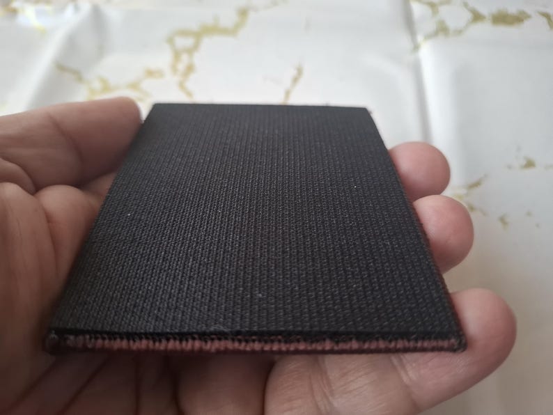 K&ouml;nnte beinhalten: Ein rechteckiger, schwarzer Stoffflicken mit strukturierter Oberfl&auml;che. Der Flicken hat einen d&uuml;nnen, r&ouml;tlich-rosa Rand. Der Artikel wird in der Hand einer Person gehalten.