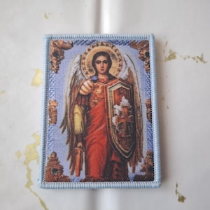 Op de afbeelding: Rechthoekige religieuze icoon met een gedetailleerde afbeelding van een engel met vleugels, die een schild en zwaard vasthoudt. De engel draagt rode en gouden gewaden tegen een blauwe achtergrond met een gouden halo. De icoon heeft een lichtblauwe rand.