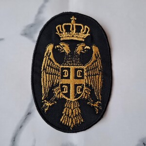 May include: Black oval patch with gold embroidered Serbian coat of arms. The design features a double-headed eagle, a crown, and a shield with a cross and four Cyrillic letters. Suitable for application on clothing or accessories.