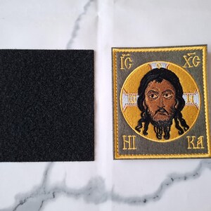 Op de afbeelding: Geborduurde patch met een religieus icoon met een gouden halo en tekst. De patch heeft een grijze rand en wordt vergezeld door een vierkant zwart klittenbandmateriaal. Het icoon toont een man met een baard.