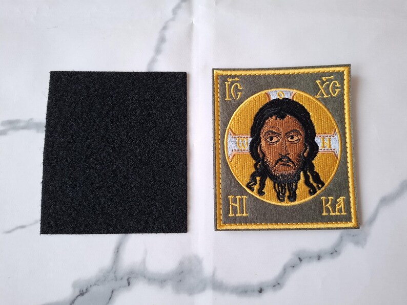 Op de afbeelding: Geborduurde patch met een religieus icoon met een gouden halo en tekst. De patch heeft een grijze rand en wordt vergezeld door een vierkant zwart klittenbandmateriaal. Het icoon toont een man met een baard.