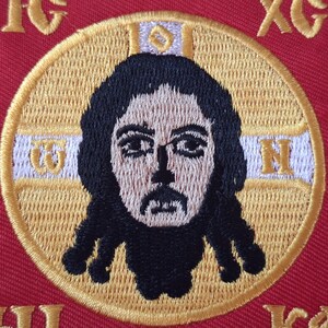 Op de afbeelding: Geborduurde patch met een gedetailleerde afbeelding van een religieuze figuur met donker haar en een baard, geplaatst in een gouden cirkel. Het ontwerp staat op een rode achtergrond met gouden letters en rand, geschikt voor religieuze kleding of ambachten.