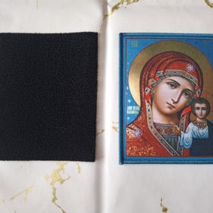 K&ouml;nnte beinhalten: Ein gestickter Aufn&auml;her mit einem religi&ouml;sen Bild einer Frau und eines Kindes mit blauem Rand. Ein separater schwarzer Quadrat aus Klettverschlussmaterial ist ebenfalls vorhanden.