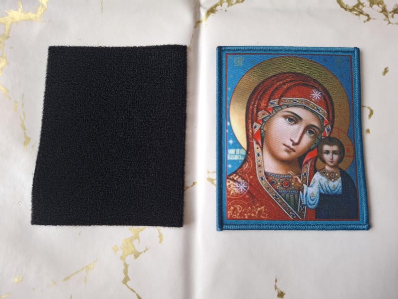 K&ouml;nnte beinhalten: Ein gestickter Aufn&auml;her mit einem religi&ouml;sen Bild einer Frau und eines Kindes mit blauem Rand. Ein separater schwarzer Quadrat aus Klettverschlussmaterial ist ebenfalls vorhanden.