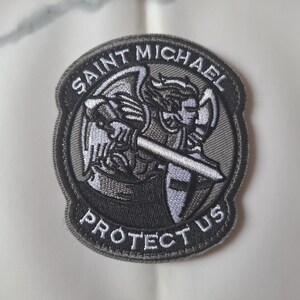 Könnte beinhalten: Runder, gestickter Aufnäher mit dem Erzengel Michael, dargestellt mit Schwert und Schild. Der Aufnäher trägt den Text "Saint Michael Protect Us" in einem kreisförmigen Muster. Das Design ist überwiegend schwarz, weiß und grau.