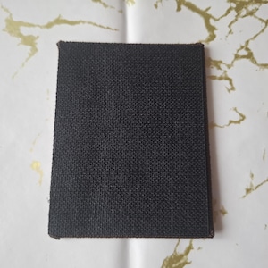 May include: A rectangular, dark brown fabric patch with a textured surface. The patch has a slightly beveled edge and is set against a white background with gold marbling.