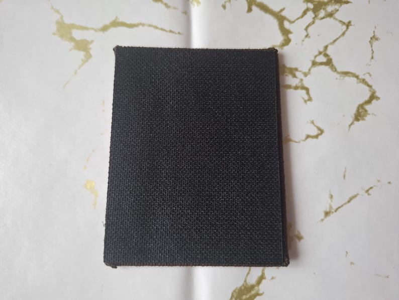 May include: A rectangular, dark brown fabric patch with a textured surface. The patch has a slightly beveled edge and is set against a white background with gold marbling.