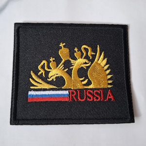 May include: A black embroidered patch with a gold double-headed eagle design and the word "RUSSIA" in red lettering. The eagle is holding a scepter and orb, and the patch features a small Russian flag.