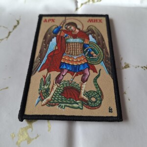 May include: Rectangular patch featuring an illustration of an angel with a sword, standing over a green dragon. The angel wears armor and a red cape. The patch has a black border and text at the top. The background is tan.