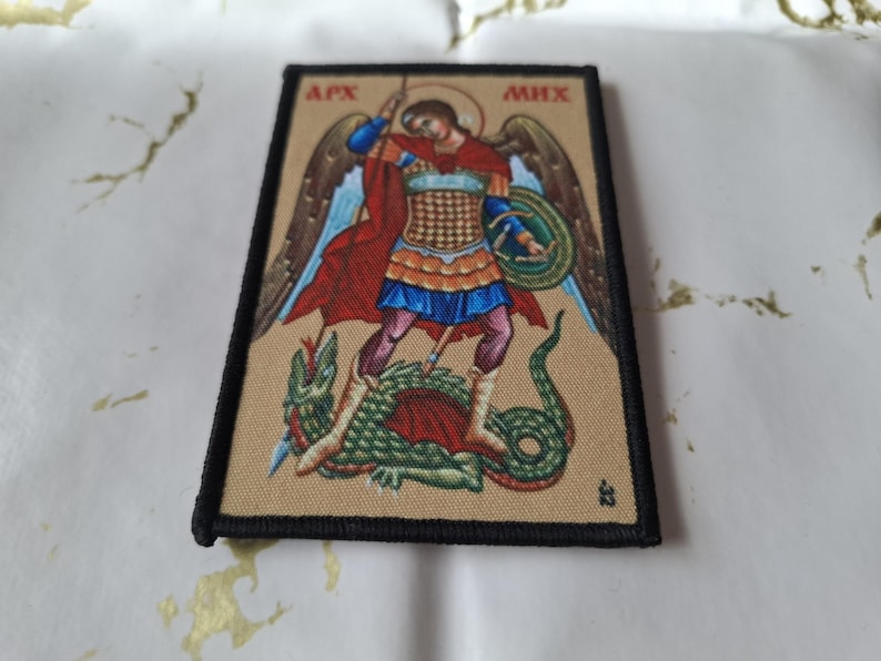 May include: Rectangular patch featuring an illustration of an angel with a sword, standing over a green dragon. The angel wears armor and a red cape. The patch has a black border and text at the top. The background is tan.