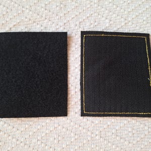 Op de afbeelding: Twee zwarte stoffen vierkanten. E&eacute;n heeft een getextureerd, gelust oppervlak en de andere is een geweven stof met gouden stiksels rond de randen. De vierkanten worden weergegeven op een wit, patroonachtig oppervlak.