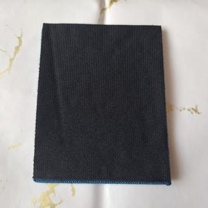 K&ouml;nnte beinhalten: Ein rechteckiger, schwarzer Stoffflicken mit gerippter Textur. Die Kanten sind sauber mit einem d&uuml;nnen blauen Rand versehen. Der Flicken befindet sich auf einem wei&szlig;en Hintergrund mit goldfarbenen Akzenten.