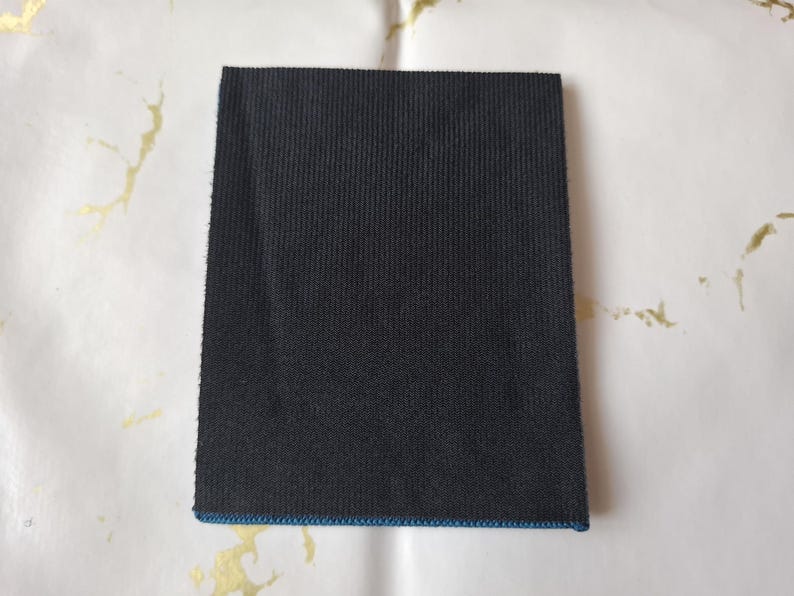 K&ouml;nnte beinhalten: Ein rechteckiger, schwarzer Stoffflicken mit gerippter Textur. Die Kanten sind sauber mit einem d&uuml;nnen blauen Rand versehen. Der Flicken befindet sich auf einem wei&szlig;en Hintergrund mit goldfarbenen Akzenten.