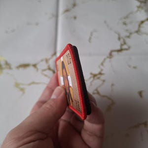 Peut inclure: &Eacute;cusson rectangulaire avec un portrait d'une personne &agrave; la peau brune et aux longs cheveux. L'&eacute;cusson a un fond beige, une bordure rouge et un dos noir. L'image est tenue par une main.