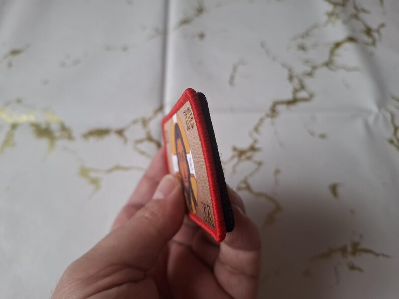 Peut inclure: &Eacute;cusson rectangulaire avec un portrait d'une personne &agrave; la peau brune et aux longs cheveux. L'&eacute;cusson a un fond beige, une bordure rouge et un dos noir. L'image est tenue par une main.