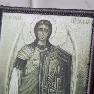 May include: Embroidered patch featuring a religious icon of an angel with a halo, holding a shield. The patch has a detailed design with muted colors and a dark border. The image includes text in a foreign language.