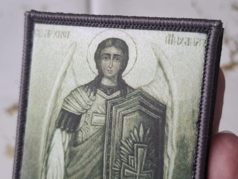 May include: Embroidered patch featuring a religious icon of an angel with a halo, holding a shield. The patch has a detailed design with muted colors and a dark border. The image includes text in a foreign language.