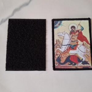 May include: A rectangular patch with a black border depicting a person on a white horse, with a red cape, spearing a dragon. Beside it is a black rectangular velcro patch.
