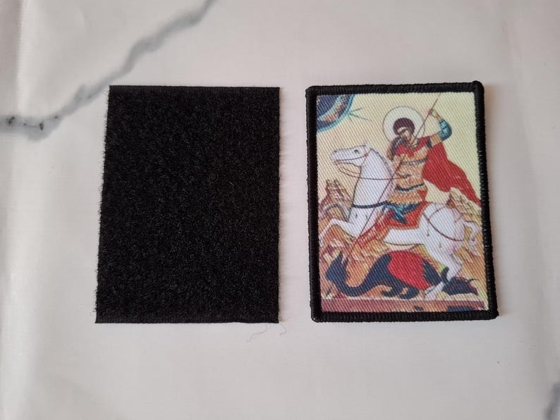 May include: A rectangular patch with a black border depicting a person on a white horse, with a red cape, spearing a dragon. Beside it is a black rectangular velcro patch.