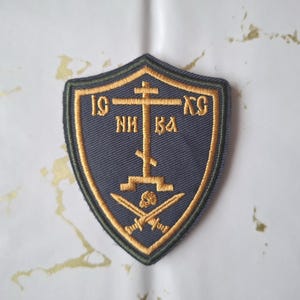 Może przedstawiać: Haftowana naszywka w kształcie tarczy, z złotym krzyżem, tekstem i skrzyżowanymi mieczami na granatowym tle. Naszywka ma złotą obwódkę i zielony kontur. Tekst zawiera litery "IC XC" i inne znaki cyrylicy.