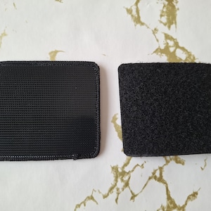 May include: Two black square pieces of fabric with a hook and loop fastener system. The fabric on the left has a woven texture, while the fabric on the right has a fuzzy texture.