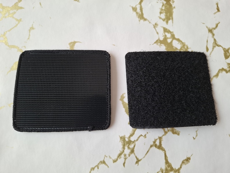 May include: Two black square pieces of fabric with a hook and loop fastener system. The fabric on the left has a woven texture, while the fabric on the right has a fuzzy texture.