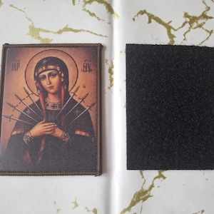 May include: A rectangular patch depicting a religious icon of a woman with a halo and swords. The image has a brown border and is next to a black, rectangular piece of fabric, likely for attachment.
