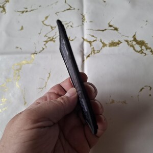 May include: A black, slim card holder with a textured surface. The card holder is held in a hand, showcasing its sleek design. The background is a white surface with gold marbling.