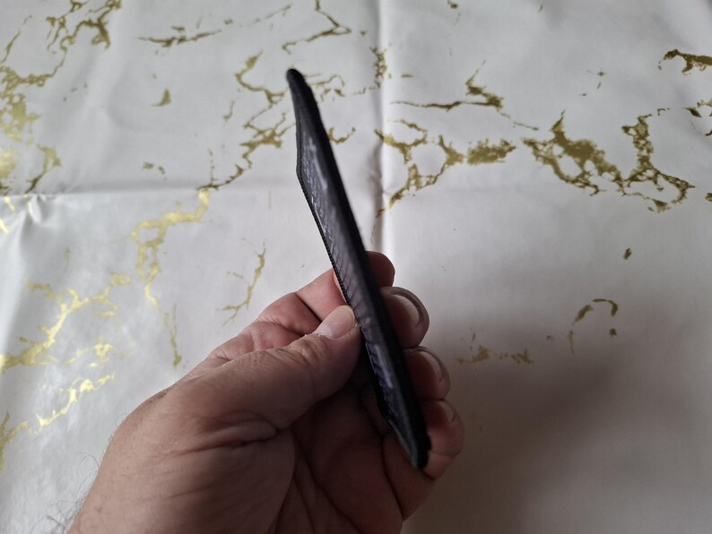 May include: A black, slim card holder with a textured surface. The card holder is held in a hand, showcasing its sleek design. The background is a white surface with gold marbling.