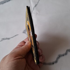 Op de afbeelding: Een rechthoekig, goudkleurig item met een zwarte rand, mogelijk een portemonnee of een kleine etui. Het item heeft een religieuze afbeelding met bruine en witte tinten. Het item wordt in de hand van een persoon gehouden.