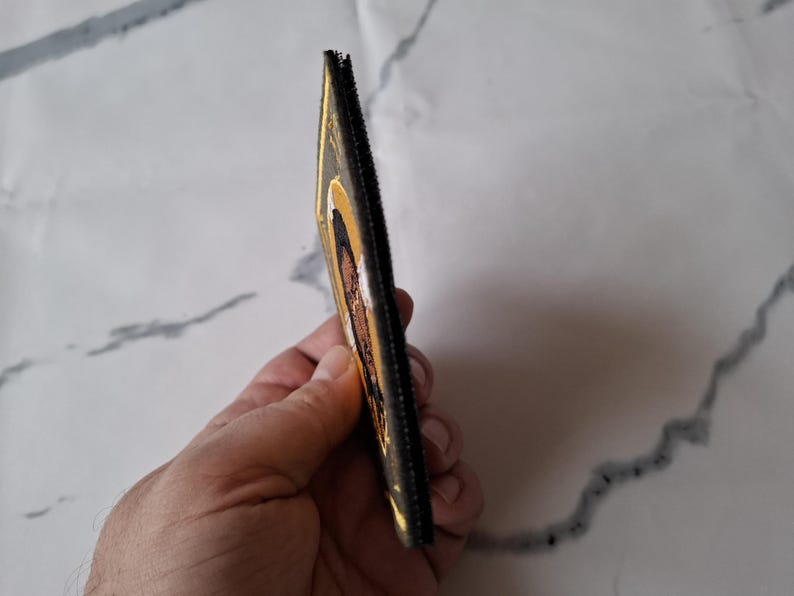 Op de afbeelding: Een rechthoekig, goudkleurig item met een zwarte rand, mogelijk een portemonnee of een kleine etui. Het item heeft een religieuze afbeelding met bruine en witte tinten. Het item wordt in de hand van een persoon gehouden.