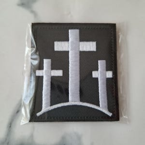 May include: A black square patch featuring three white crosses of varying sizes, arranged on a curved line. The crosses are embroidered with white thread, and the patch is encased in clear plastic.
