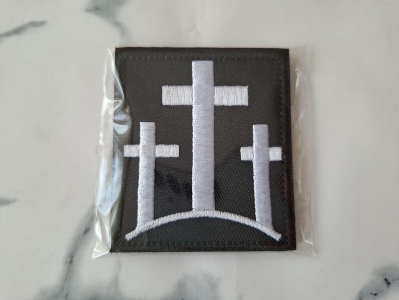 May include: A black square patch featuring three white crosses of varying sizes, arranged on a curved line. The crosses are embroidered with white thread, and the patch is encased in clear plastic.
