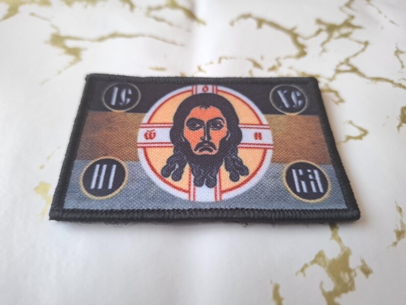 Peut inclure: &Eacute;cusson rectangulaire avec une ic&ocirc;ne religieuse. Il pr&eacute;sente une image centrale d'un homme barbu dans un cadre circulaire, avec du texte et des symboles en forme de croix. Le fond est marron et gris, avec une bordure noire.