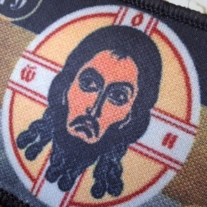 Peut inclure: Un &eacute;cusson rectangulaire repr&eacute;sentant une ic&ocirc;ne religieuse. L'image centrale est une repr&eacute;sentation stylis&eacute;e d'un homme barbu, dans un cadre circulaire avec un motif de croix. La palette de couleurs comprend l'orange, le noir et le beige.