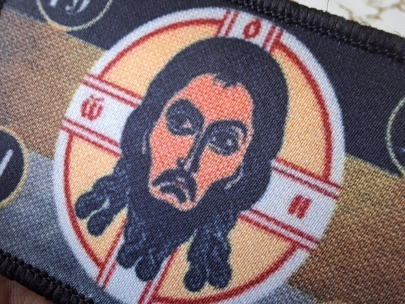 Peut inclure: Un &eacute;cusson rectangulaire repr&eacute;sentant une ic&ocirc;ne religieuse. L'image centrale est une repr&eacute;sentation stylis&eacute;e d'un homme barbu, dans un cadre circulaire avec un motif de croix. La palette de couleurs comprend l'orange, le noir et le beige.