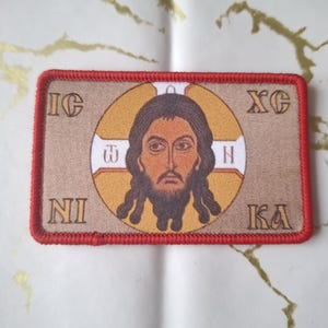 Może przedstawiać: Prostokątna naszywka z czerwoną obwódką przedstawiająca ikonę religijną. Ikona przedstawia mężczyznę z długimi brązowymi włosami i brodą w złotym okręgu. Greckie litery są widoczne wokół okręgu.