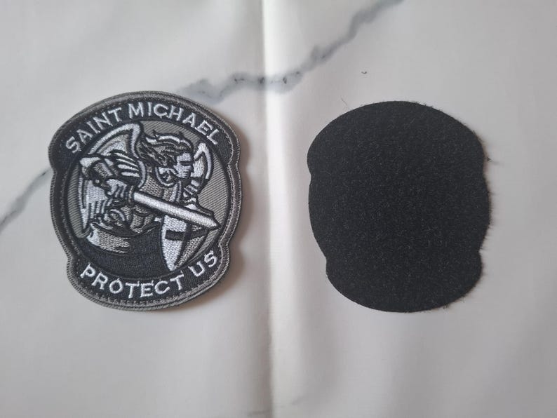 May include: Embroidered patch depicting Saint Michael with a sword and shield, with the text "SAINT MICHAEL PROTECT US" in a circular design. A separate black felt patch is also visible.