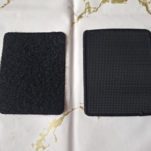 May include: Two black rectangular hook and loop fastener patches. One has a fuzzy texture, the other a woven, grid-like texture. The patches are on a white surface with gold accents.