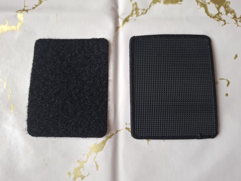 May include: Two black rectangular hook and loop fastener patches. One has a fuzzy texture, the other a woven, grid-like texture. The patches are on a white surface with gold accents.
