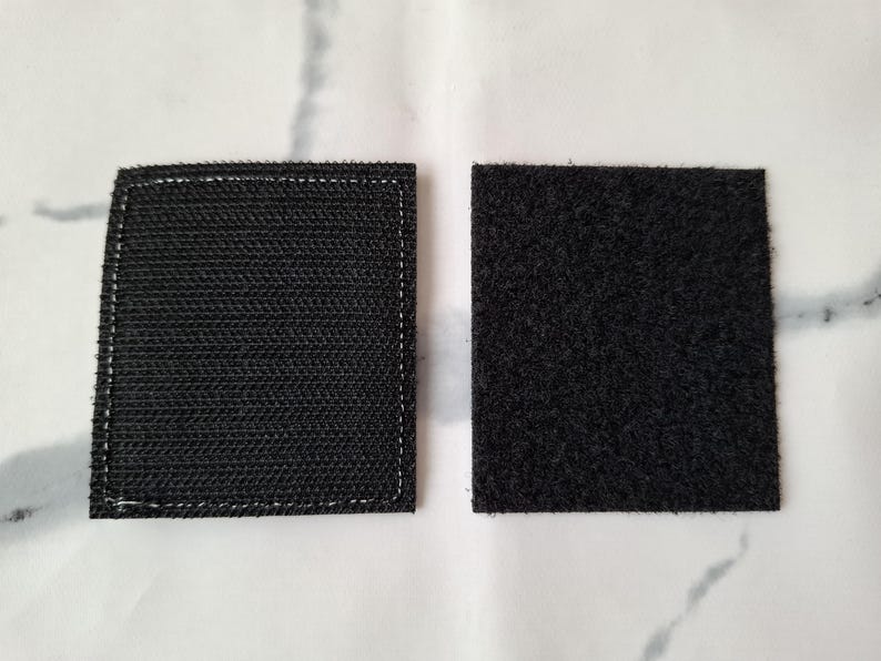 May include: Two black square hook and loop fastener patches. One patch has a woven texture with white stitching around the edges. The other patch has a fuzzy texture.