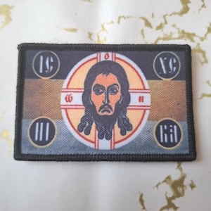 May include: Rectangular patch with a religious icon. It features a central image of a bearded man within a cross and halo. The patch has a black border and circular text elements in each corner. The color palette includes shades of brown, orange, and blue.