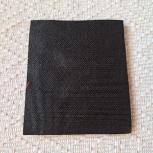 Op de afbeelding: Een vierkante, zwarte stoffen patch met een getextureerd oppervlak. De patch is geplaatst tegen een witte, patroonachtergrond. Een kleine, rode draad is zichtbaar op een rand.