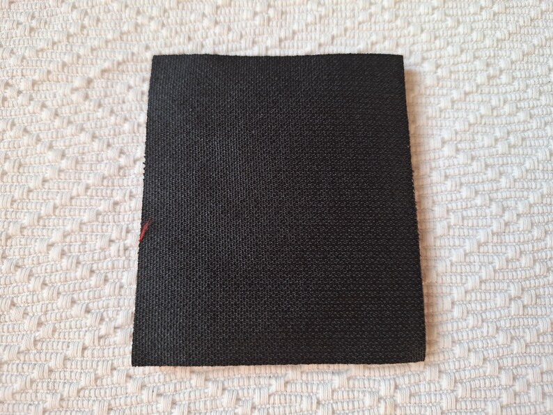 Op de afbeelding: Een vierkante, zwarte stoffen patch met een getextureerd oppervlak. De patch is geplaatst tegen een witte, patroonachtergrond. Een kleine, rode draad is zichtbaar op een rand.