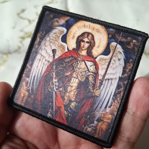 May include: Square patch featuring an illustration of an angel with a halo, wings, and a sword. The angel is depicted in armor and a red cape. The patch has a black border and is held in a hand.