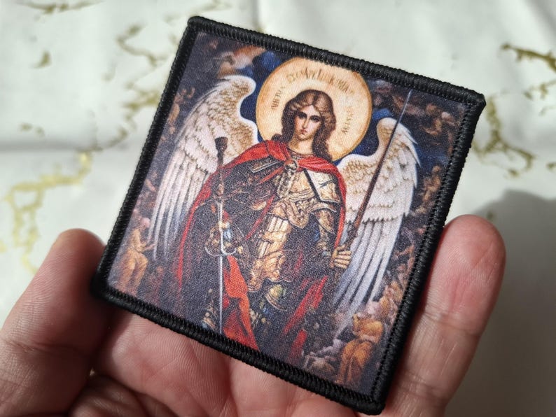 May include: Square patch featuring an illustration of an angel with a halo, wings, and a sword. The angel is depicted in armor and a red cape. The patch has a black border and is held in a hand.