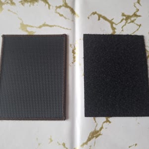 May include: Two square patches, one with a dark grey, textured surface and brown stitching, and the other with a black, fuzzy surface. The patches are displayed on a white background with gold accents.